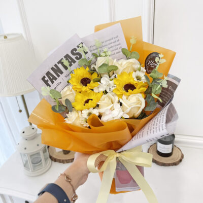 a flower bouquet of soap sunflowers and champagne soap roses wrapped in orange and newspaper-style paper with a pale yellow ribbon