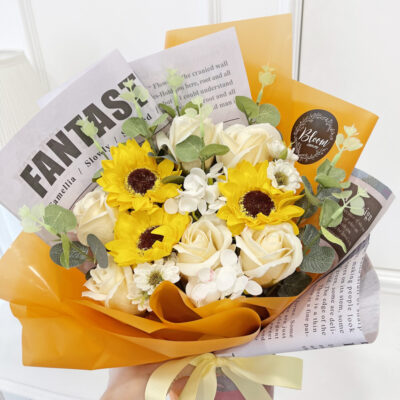 wrapped in orange translucent paper, this soap flower bouquet features bright sunflowers and soft champagne roses with green leaves
