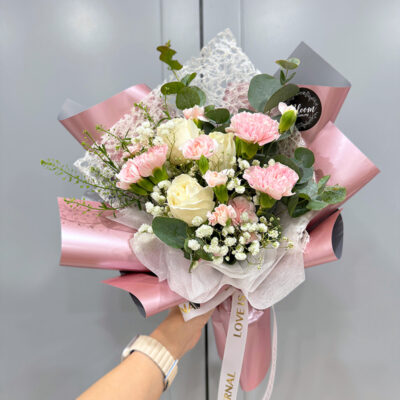 a flower bouquet wrapped in pink paper featuring white roses, pink carnations, pink carnation spray, and baby's breath with greenery